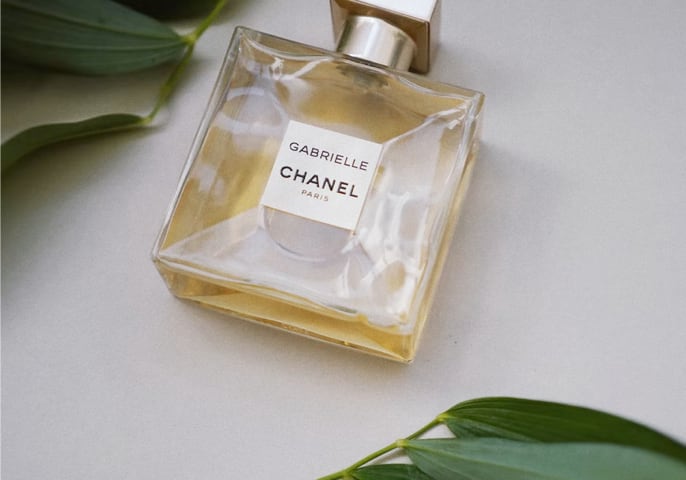 A bottle of Gabrielle Chanel perfume laying on a white background surrounded by fern leaves